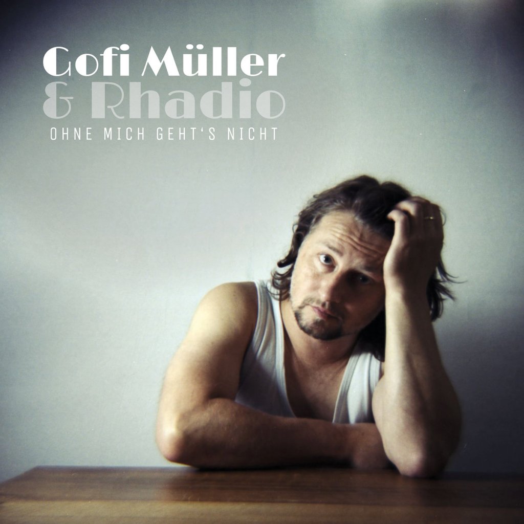 Portrait of a man in a white tank top resting his chin on his hand while sitting at a table, expressing a thoughtful or contemplative mood. The background is neutral and soft-focused. The text 'Gofi Müller & Rhadio' and the title 'Ohne mich geht's nicht' are prominently displayed.